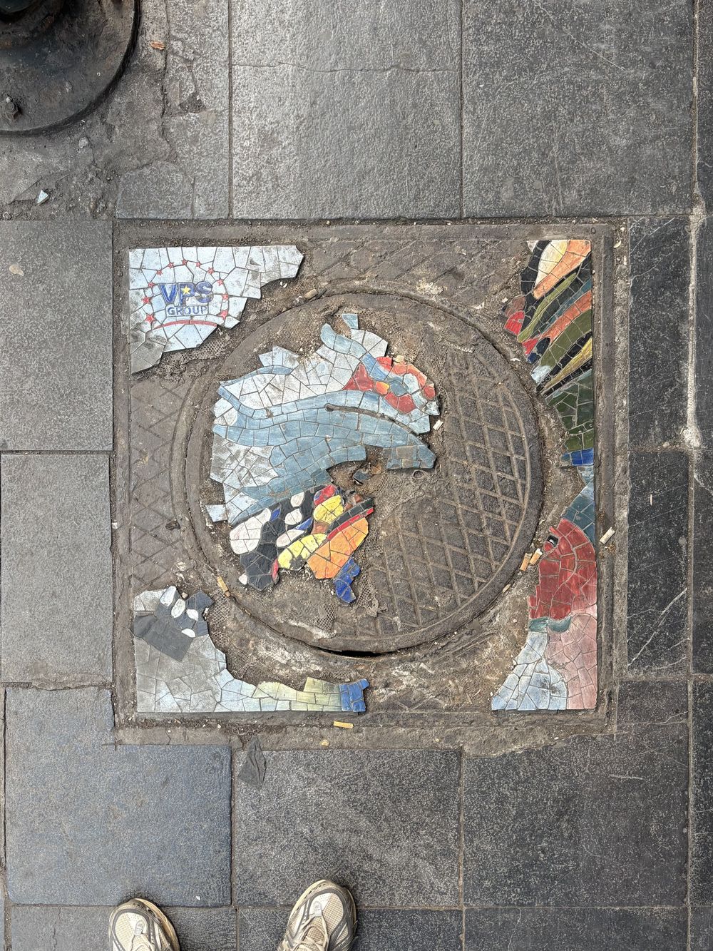 Broken colorful mosaic on a manhole cover in a stone sidewalk