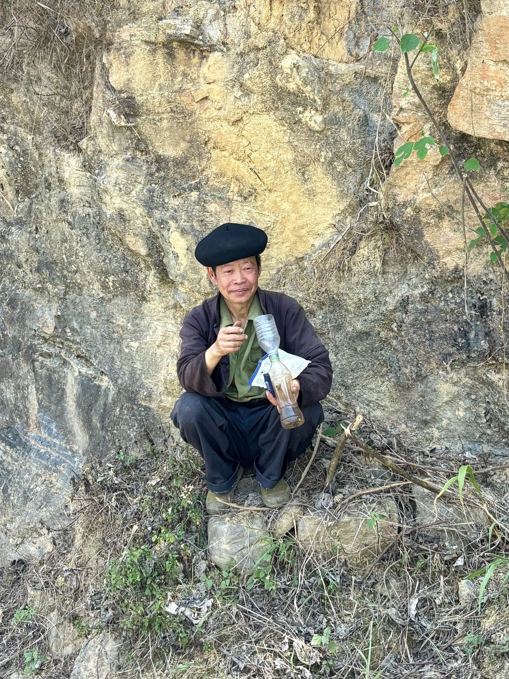 Man in a black beret crouching against a rock wall, holding a small bottle and a wrapped item